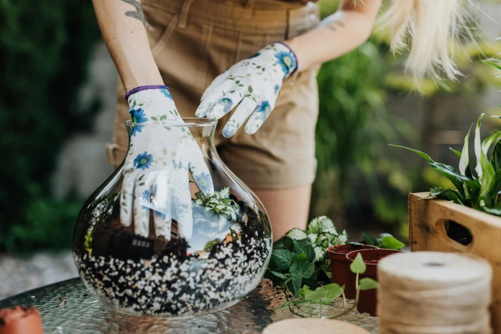 A hand carefully adding potting mix on top of a layer of pebbles and charcoal inside a glass terrarium, showing the step-by-step process of building a healthy foundation for plant growth.